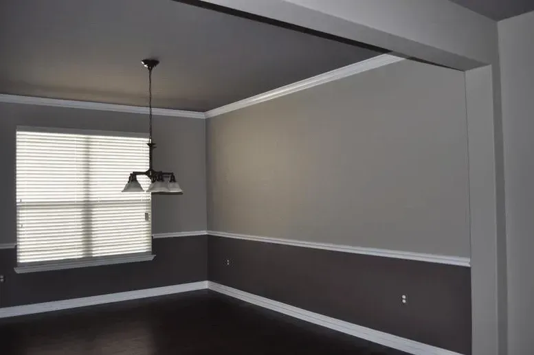 Empty dining room with gray walls, white trim, dark floor, and a hanging light fixture near a window.