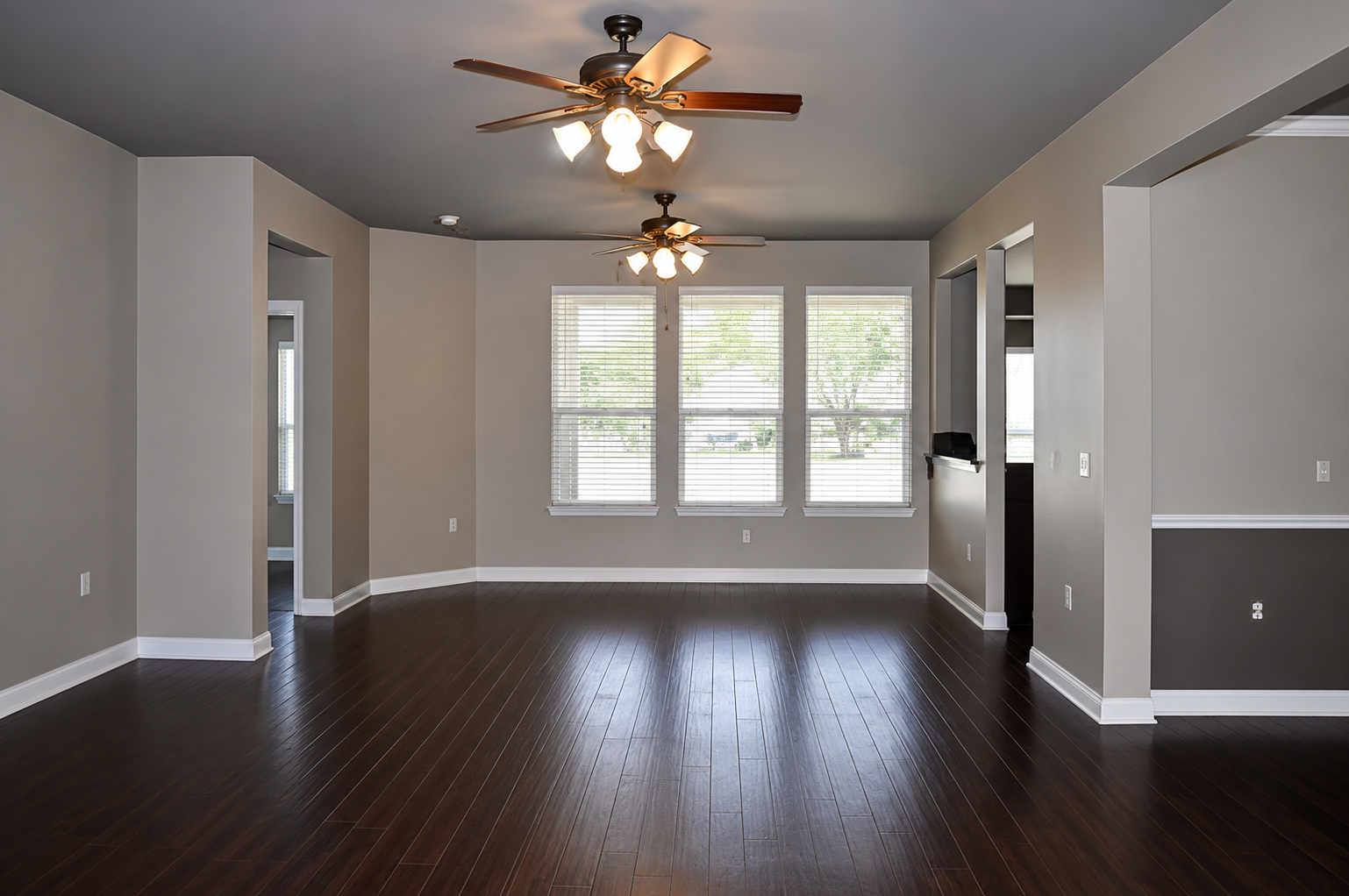 Empty modern living room with dark wood floors, gray walls, ceiling fans, and bright front windows