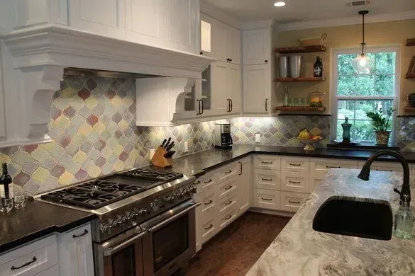 Modern white kitchen with colorful tile backsplash, stainless stove, marble counters, and window over sink