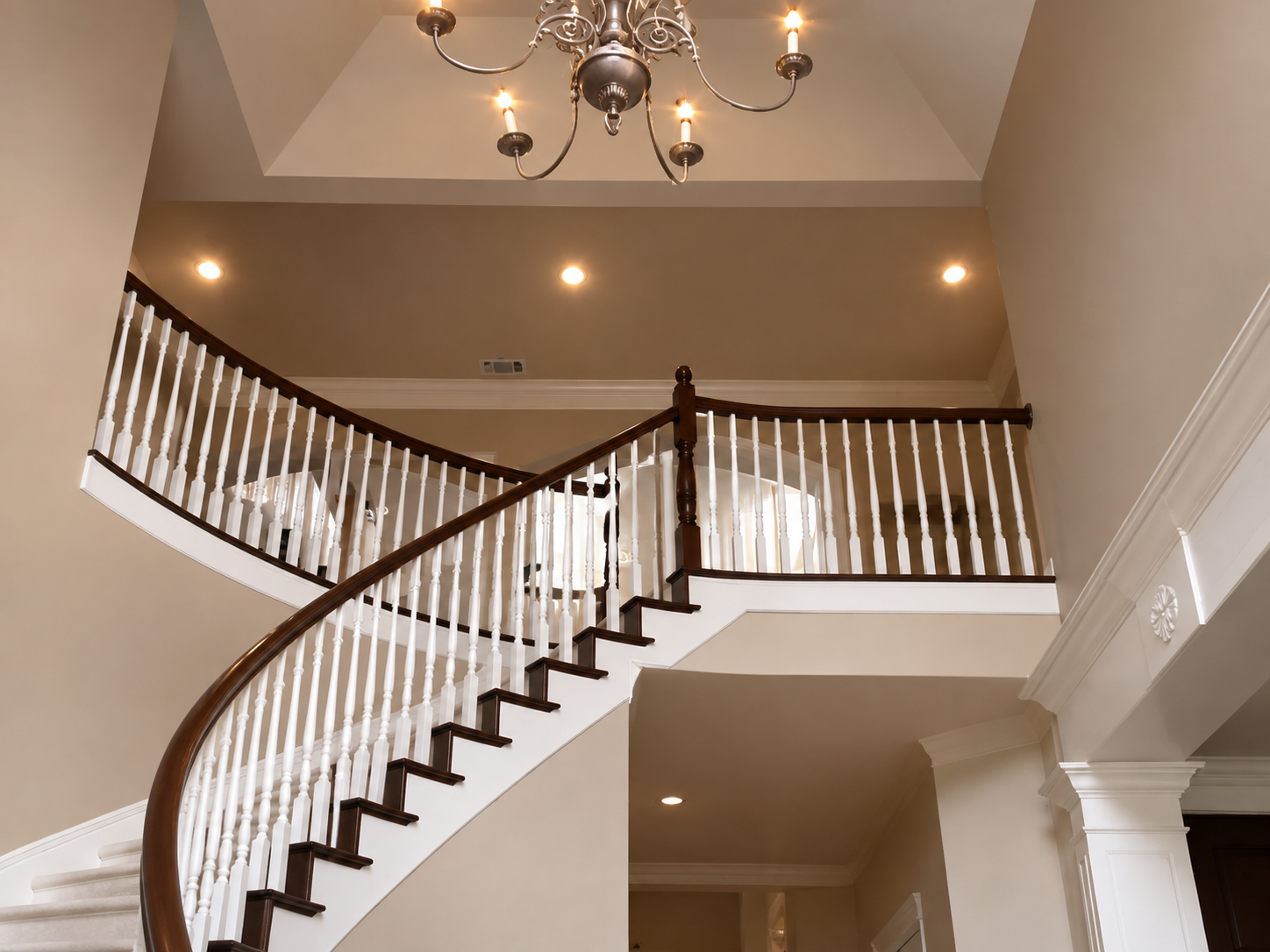 Curved staircase with white balusters and dark railing under a chandelier in a beige foyer