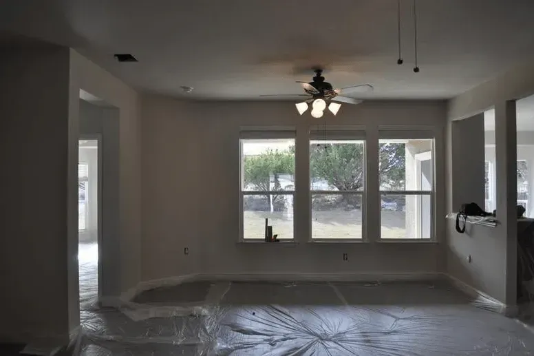Empty living room with plastic-covered floor, ceiling fan, and large windows overlooking a fenced yard