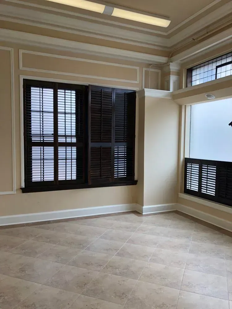 Empty beige room with tiled floor, large dark window blinds, and a high transom window.