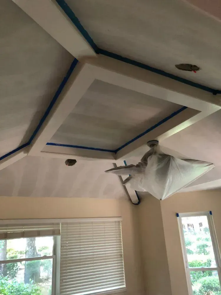 Ceiling being painted white with blue tape, while a covered light fixture hangs in a room with windows.