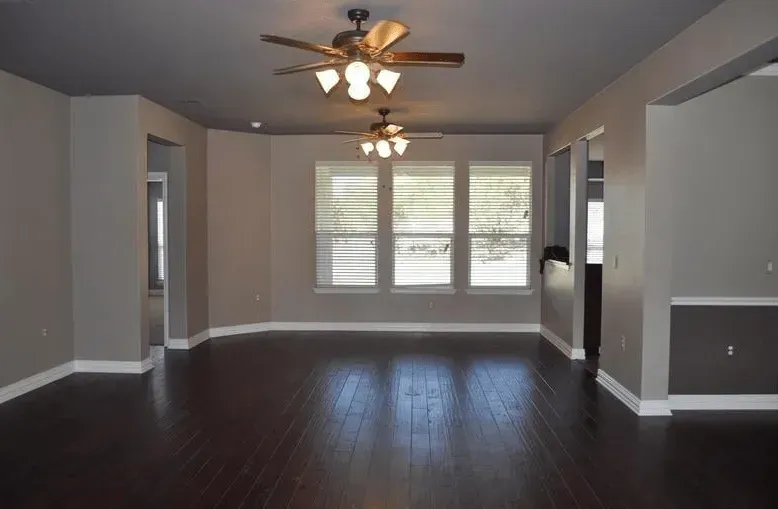Empty living room with dark wood floors, gray walls, ceiling fans, and windows with blinds