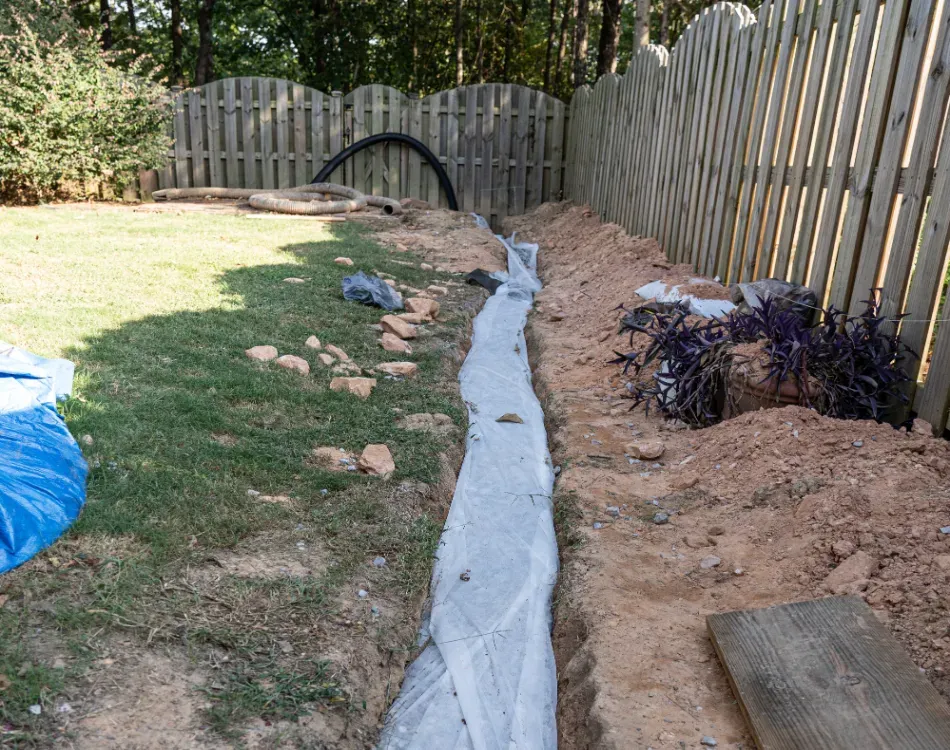 A drainage system is being installed in a backyard next to a wooden fence.