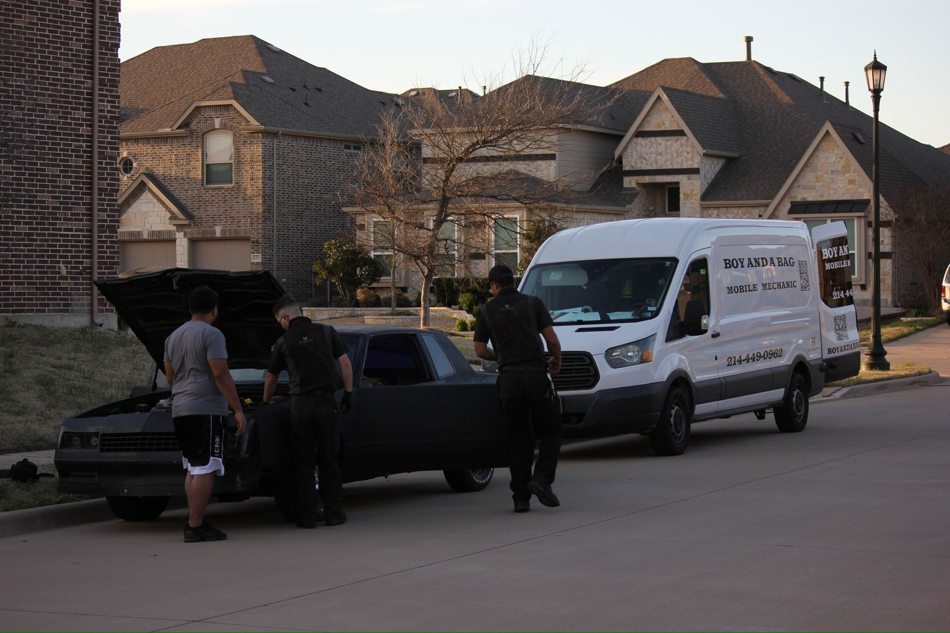 People loading items into a black car beside a white van on a suburban street at dusk