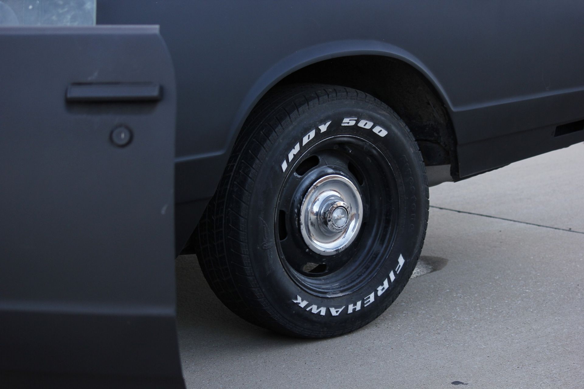 Close-up of a black vehicle’s rear tire and wheel on pavement, with a dark side panel visible.