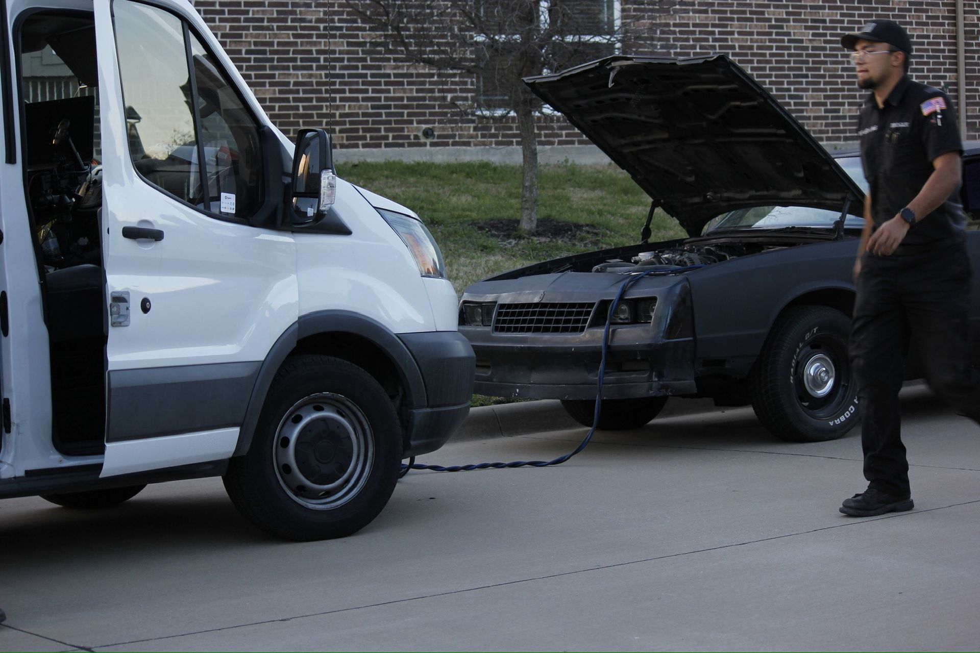 Black car with hood open and white van parked nearby; a man stands beside them on a street.