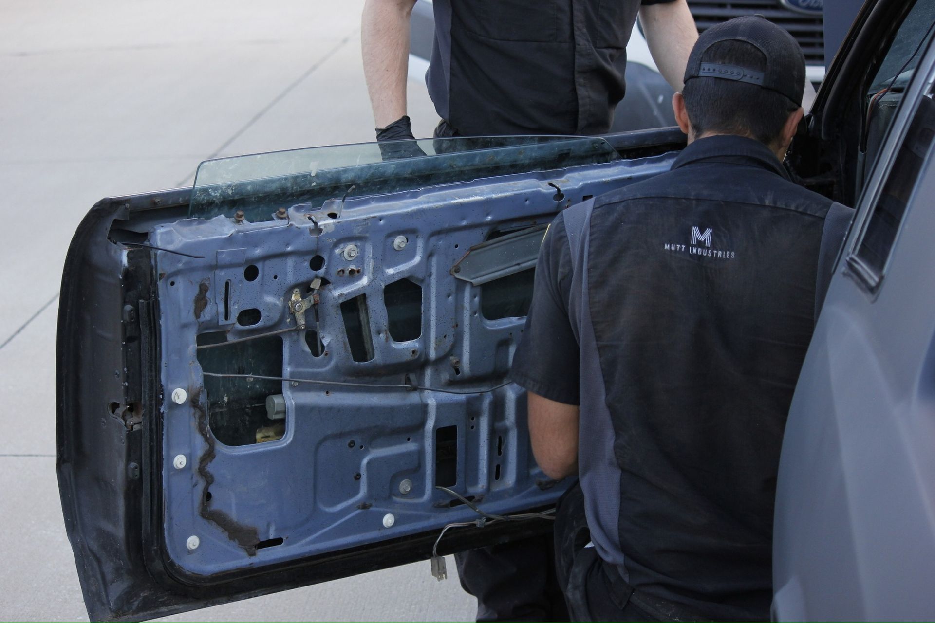Two workers removing a car door panel in a garage, exposing the interior of the door.