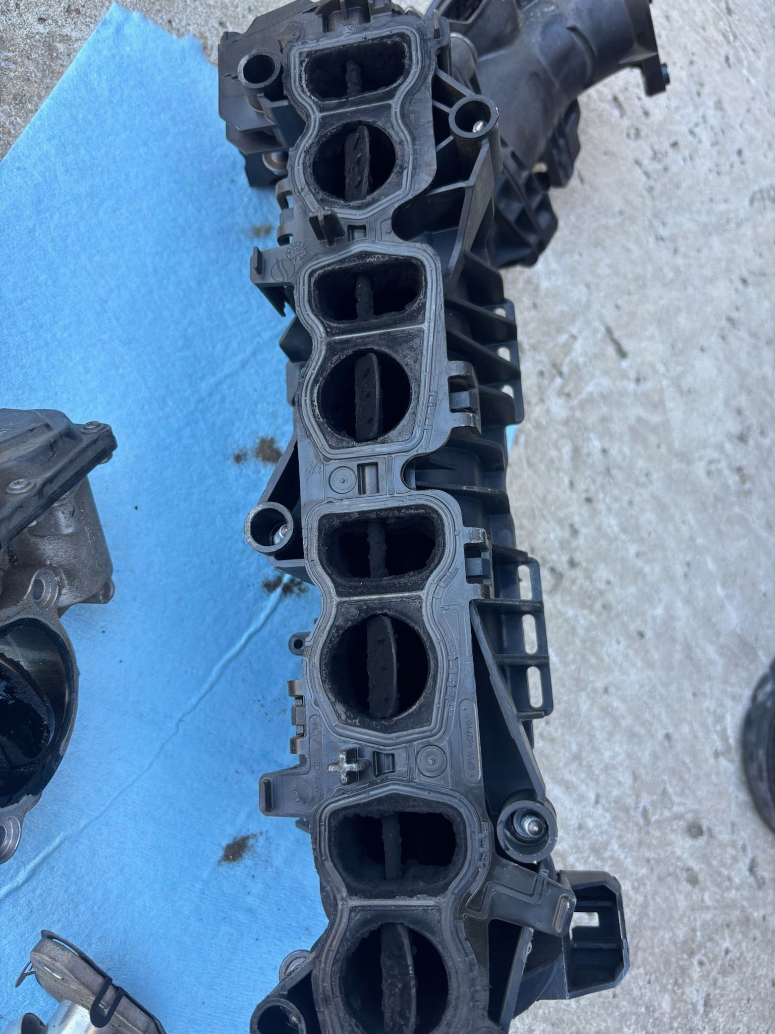Black intake manifold on a blue mat beside another car part on concrete ground