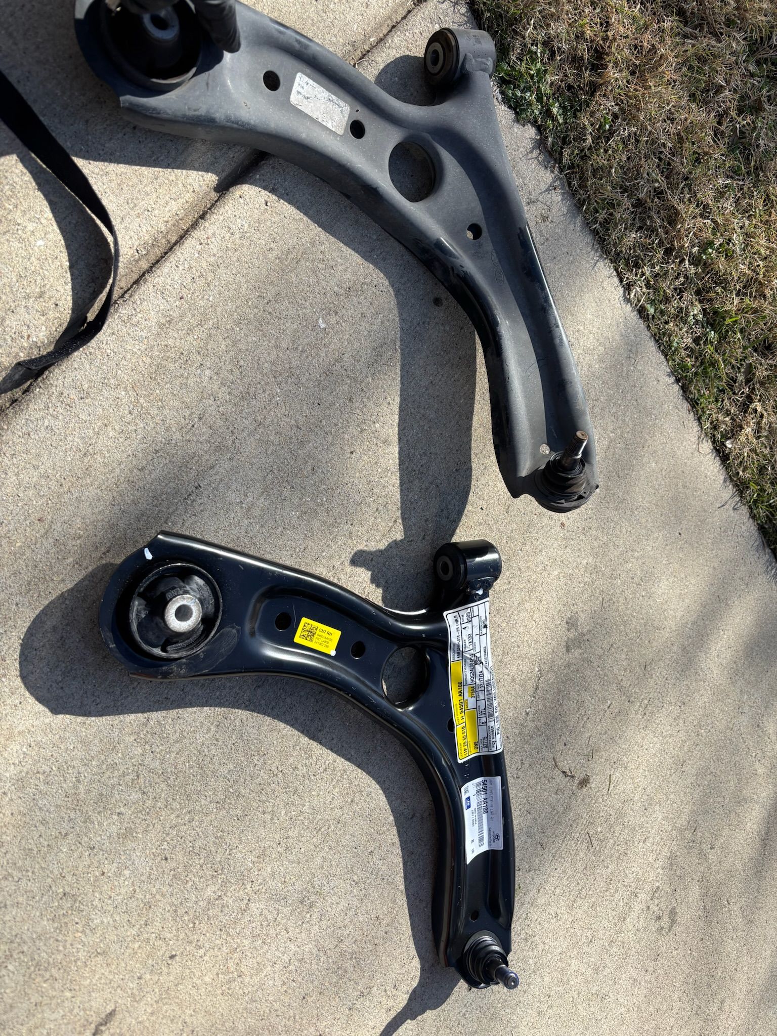 Two black metal suspension control arms lying on concrete next to grass