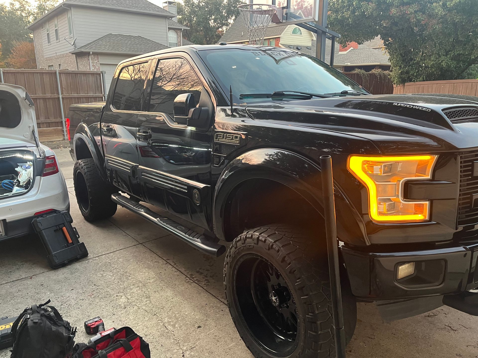 Black lifted pickup truck parked in a driveway, with amber headlights on and a white car beside it.
