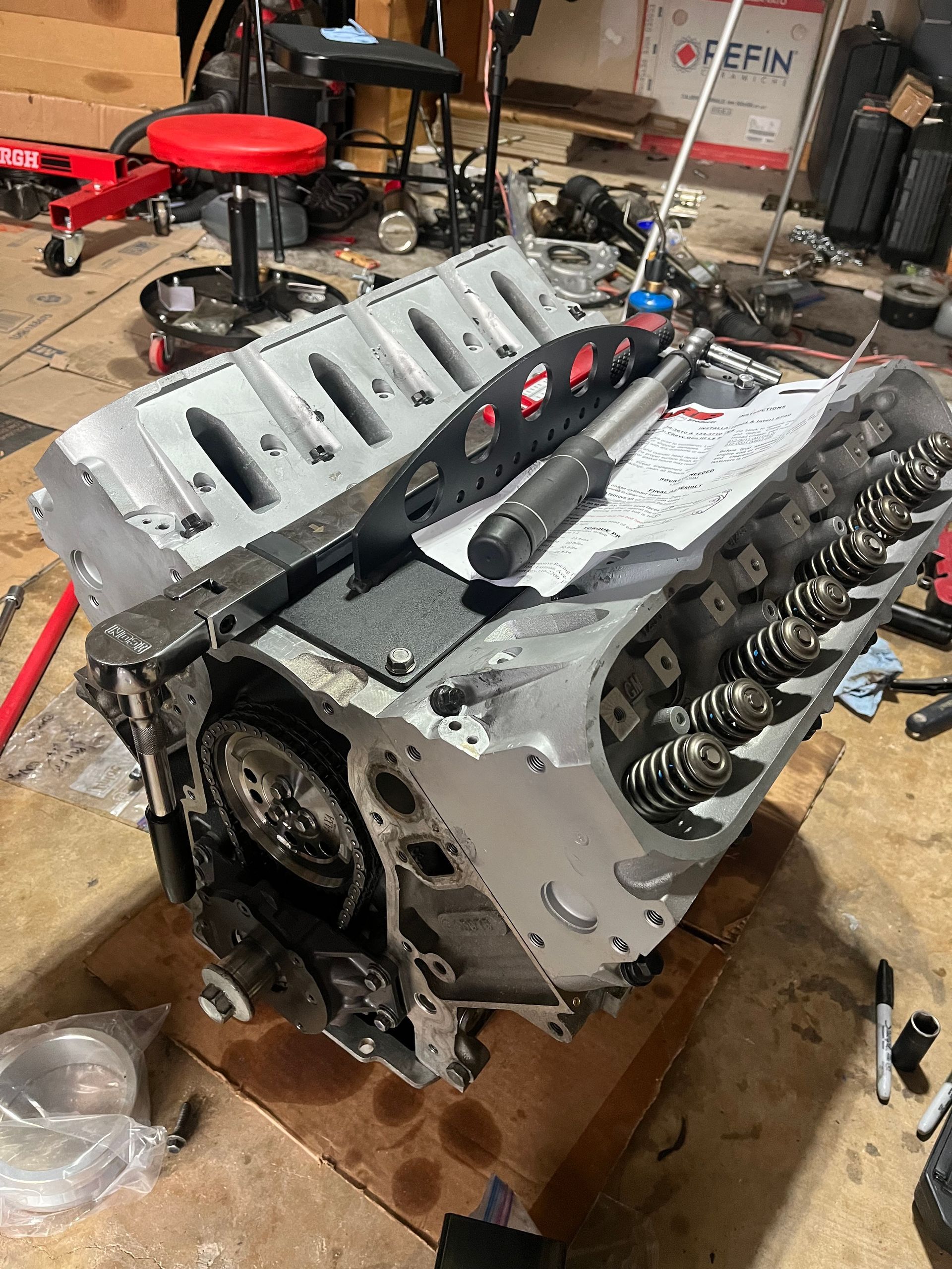 Partially disassembled gray V8 engine block on a workshop floor with tools and parts nearby