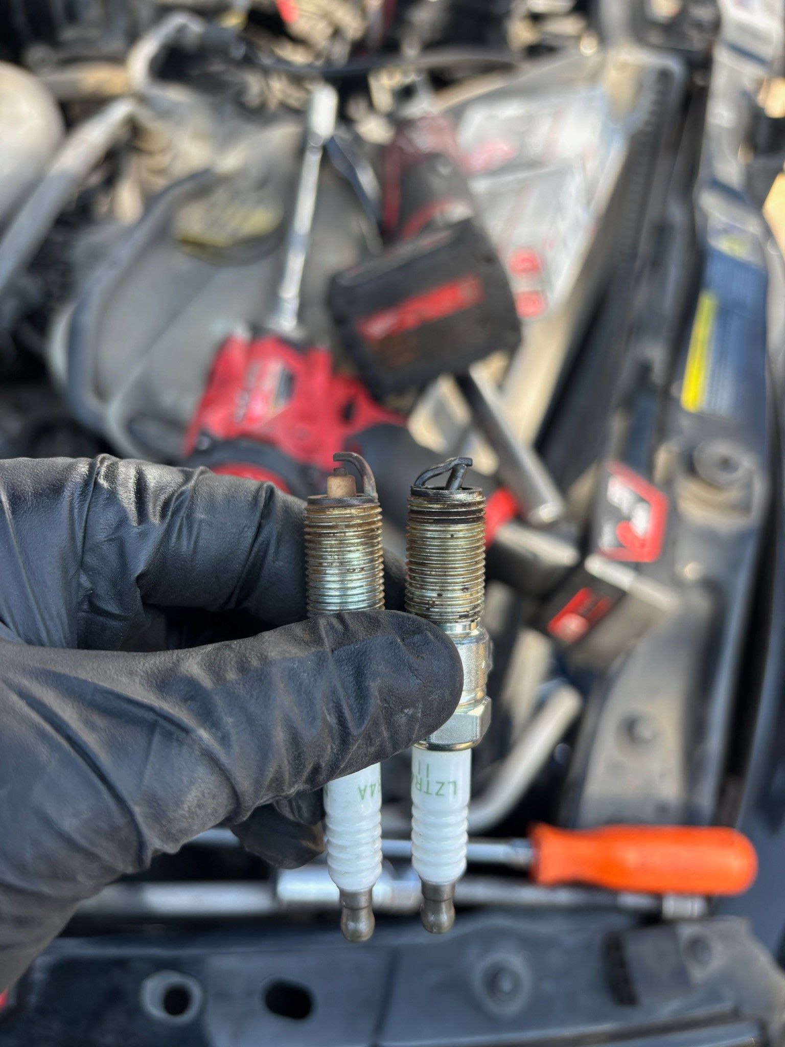 Gloved hand holding two worn spark plugs in front of a blurred engine bay