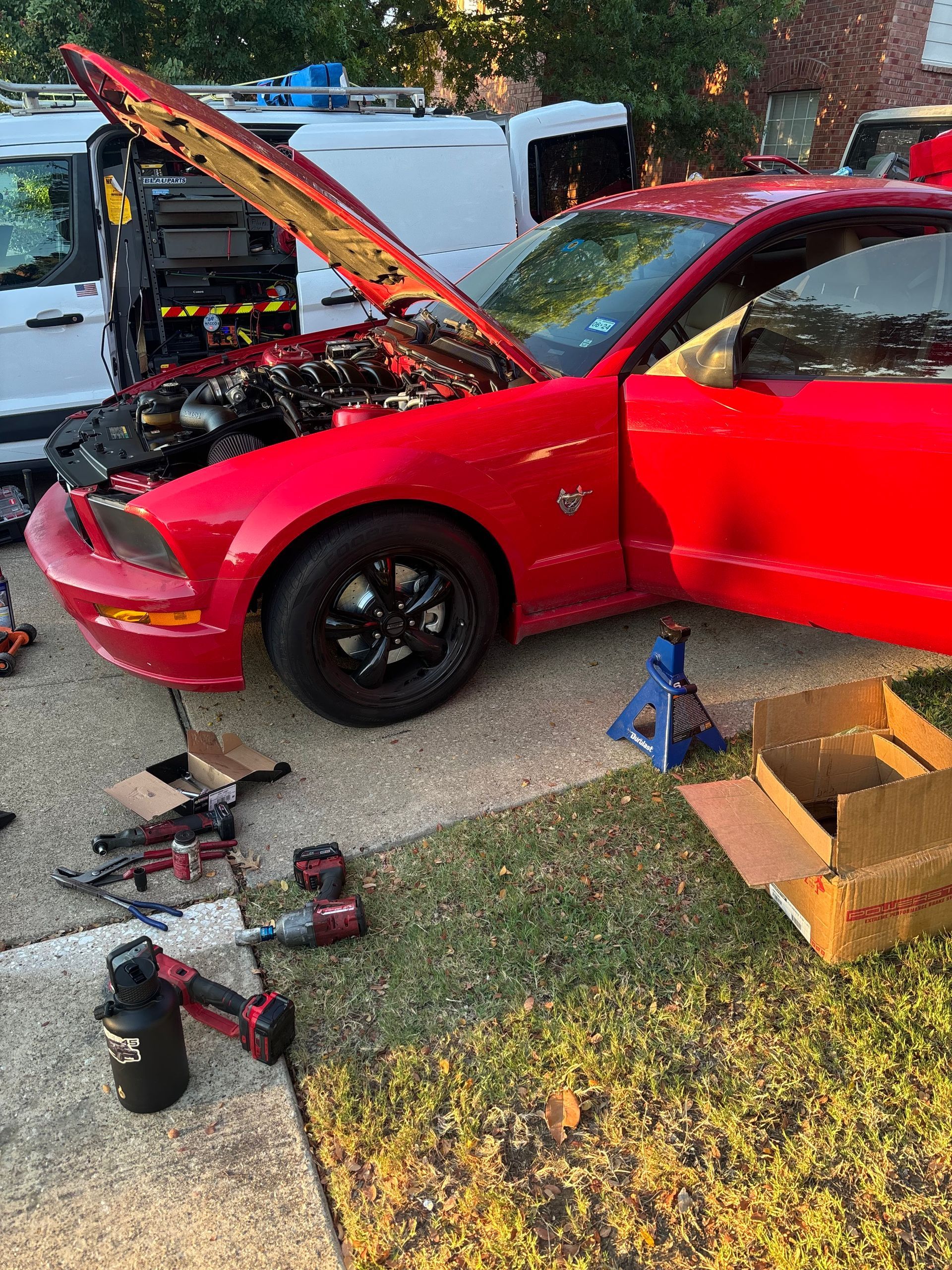 Red car with hood open in a driveway, surrounded by tools and parts, with a white truck parked behind it.