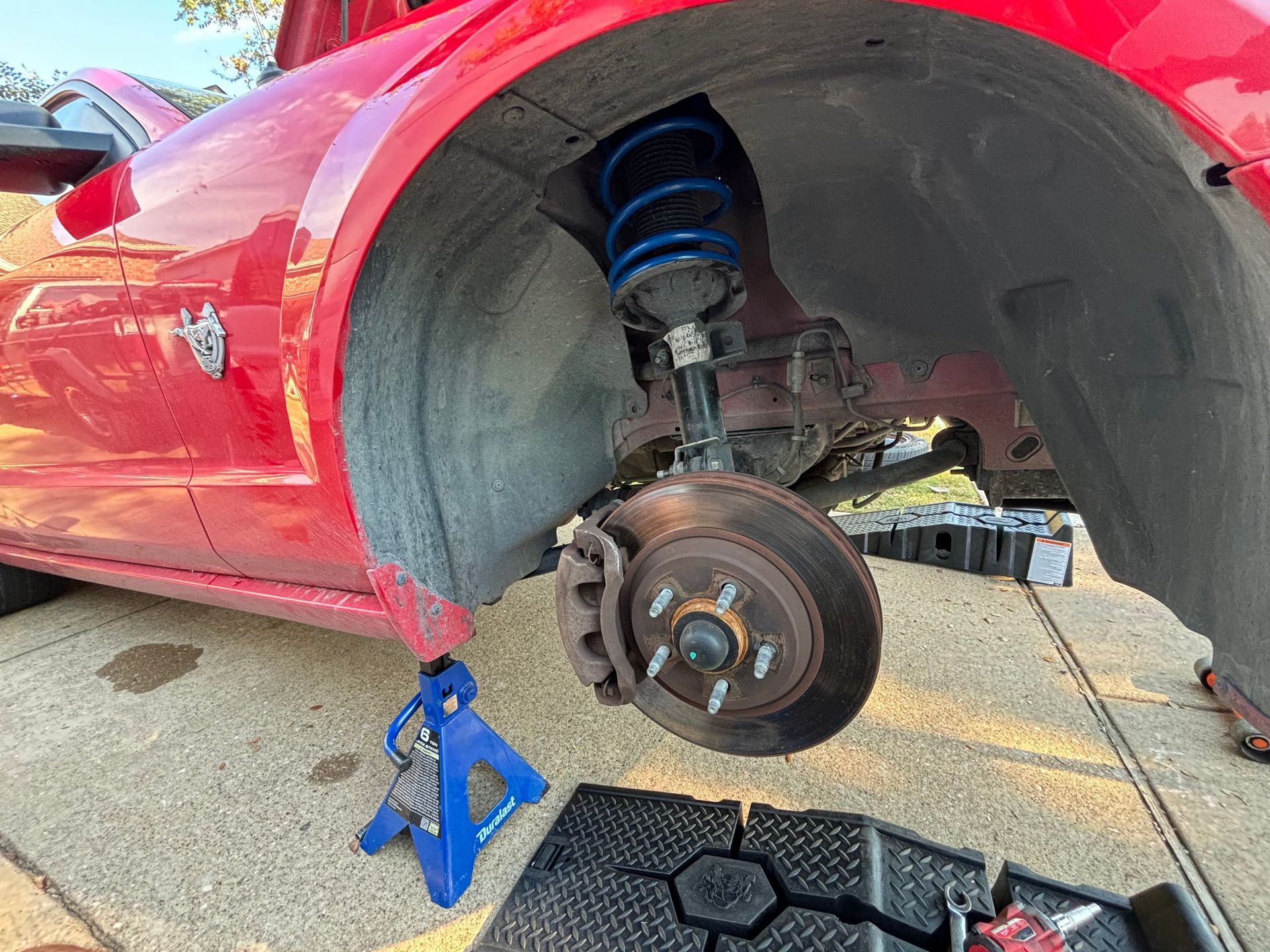 Red sports car on jack stands with front wheel removed, exposing brake and suspension parts.