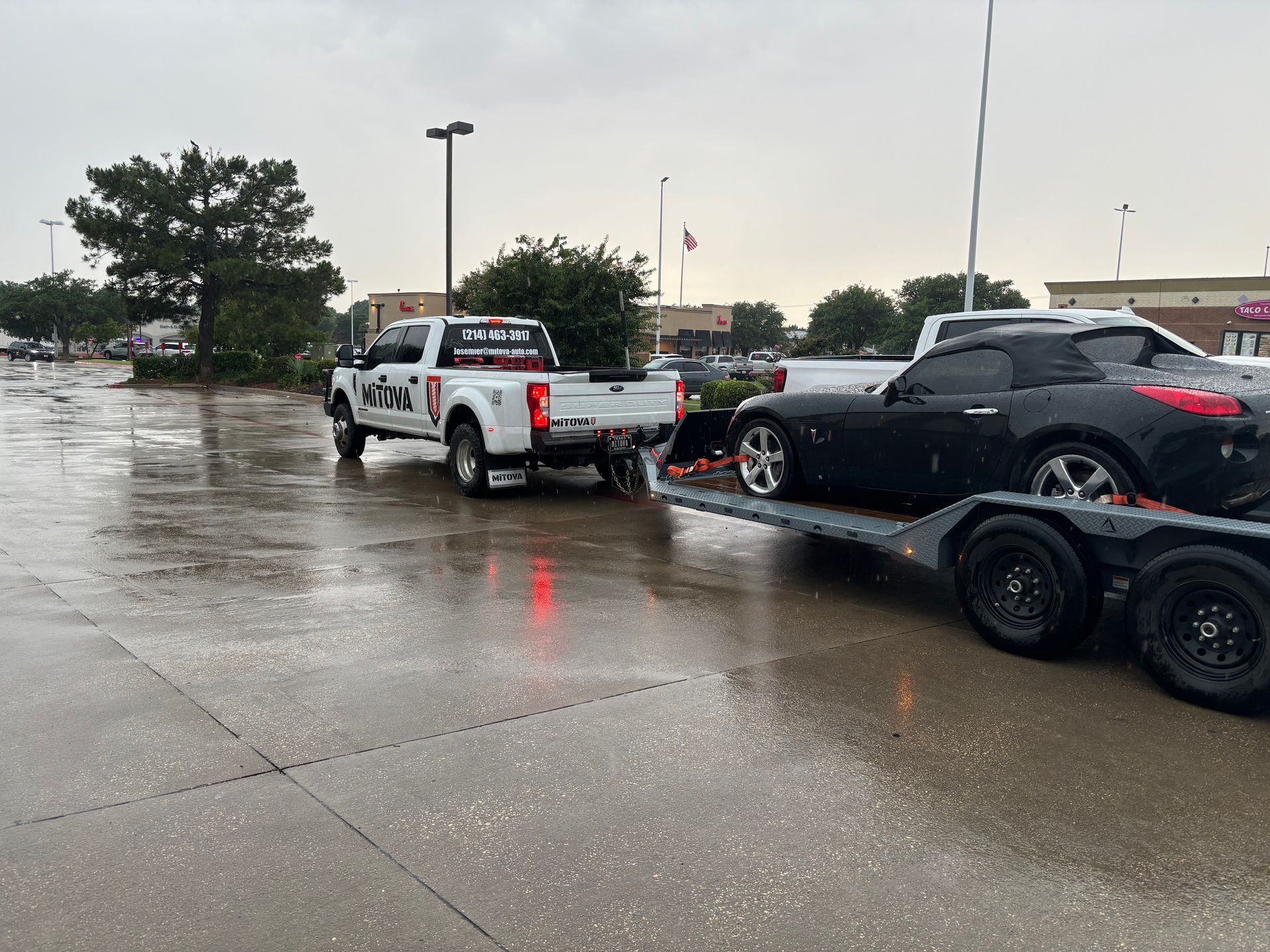 Tow truck towing a black SUV in a rainy parking lot, with a white pickup nearby.