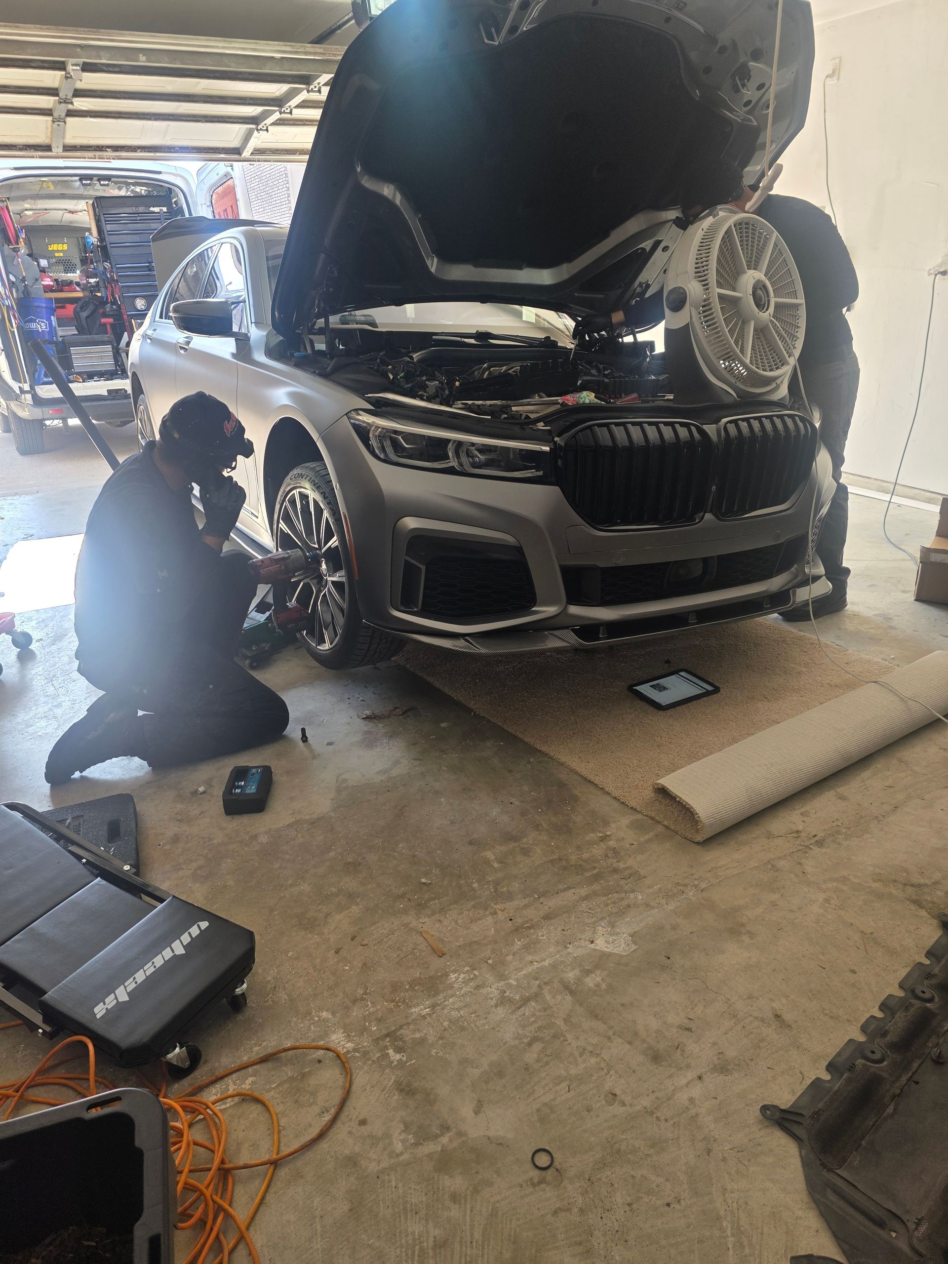 Black SUV in a garage with hood open and front end damage; tools and a mechanic nearby.