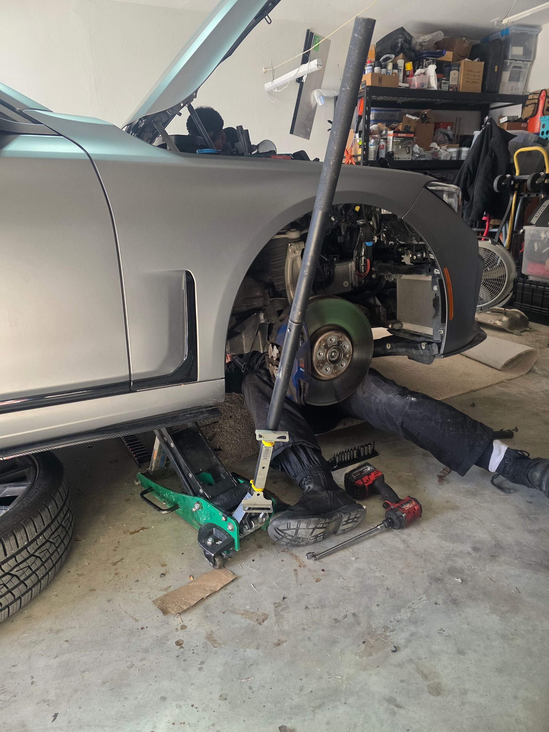Silver car jacked up in a garage, with a person working on the front wheel area and tools nearby