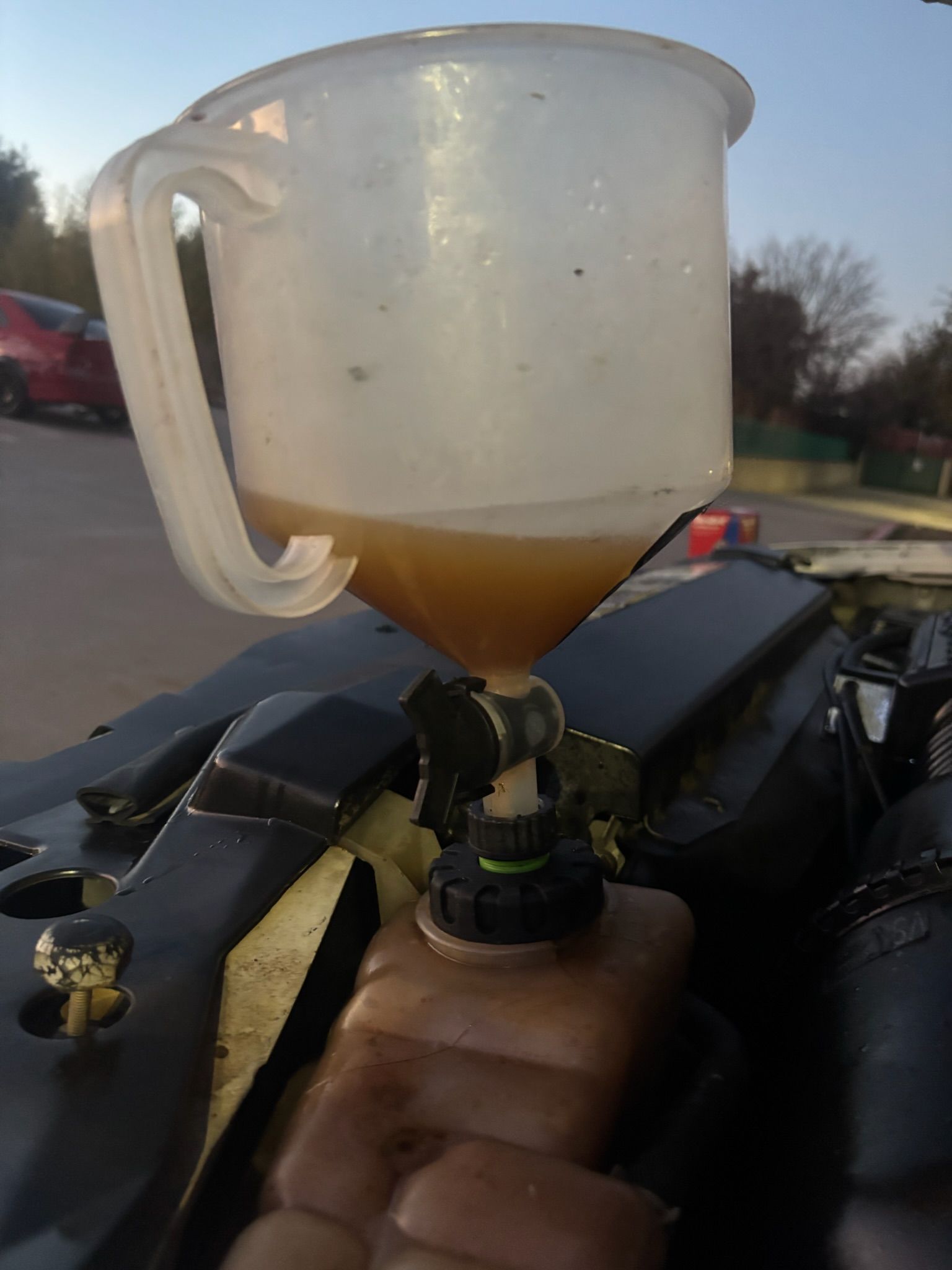 Translucent plastic funnel cup with amber liquid, held above a black engine part outdoors at sunset