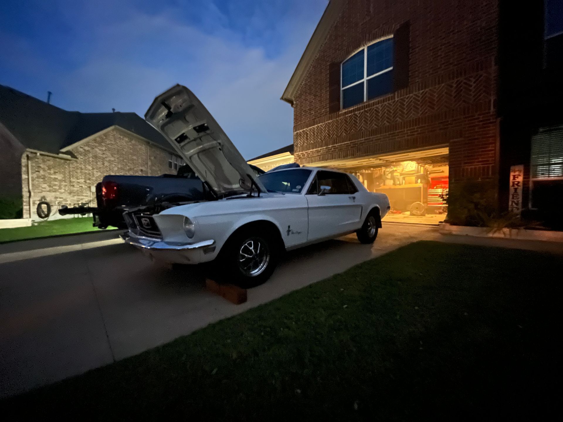 Classic white car with hood open in a driveway at dusk beside a lit brick house