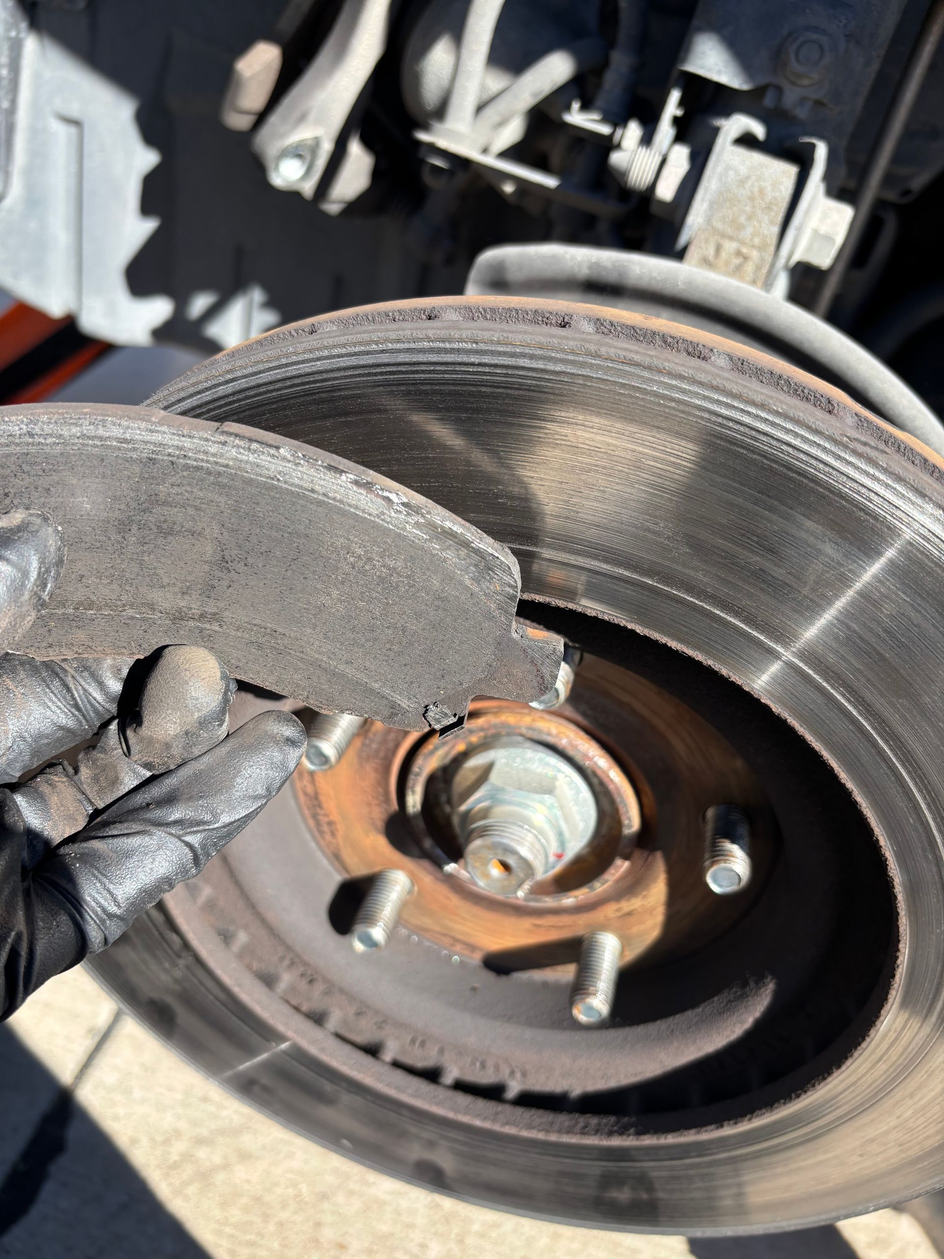 Close-up of a worn car brake rotor and caliper with a gloved hand pointing at the pad.