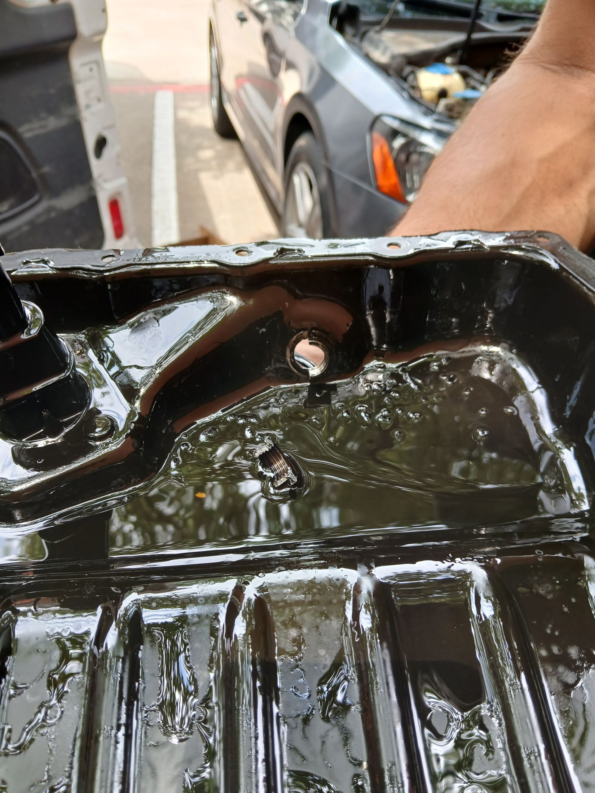Close-up of a black oil pan filled with dark liquid, with a car visible in the background.