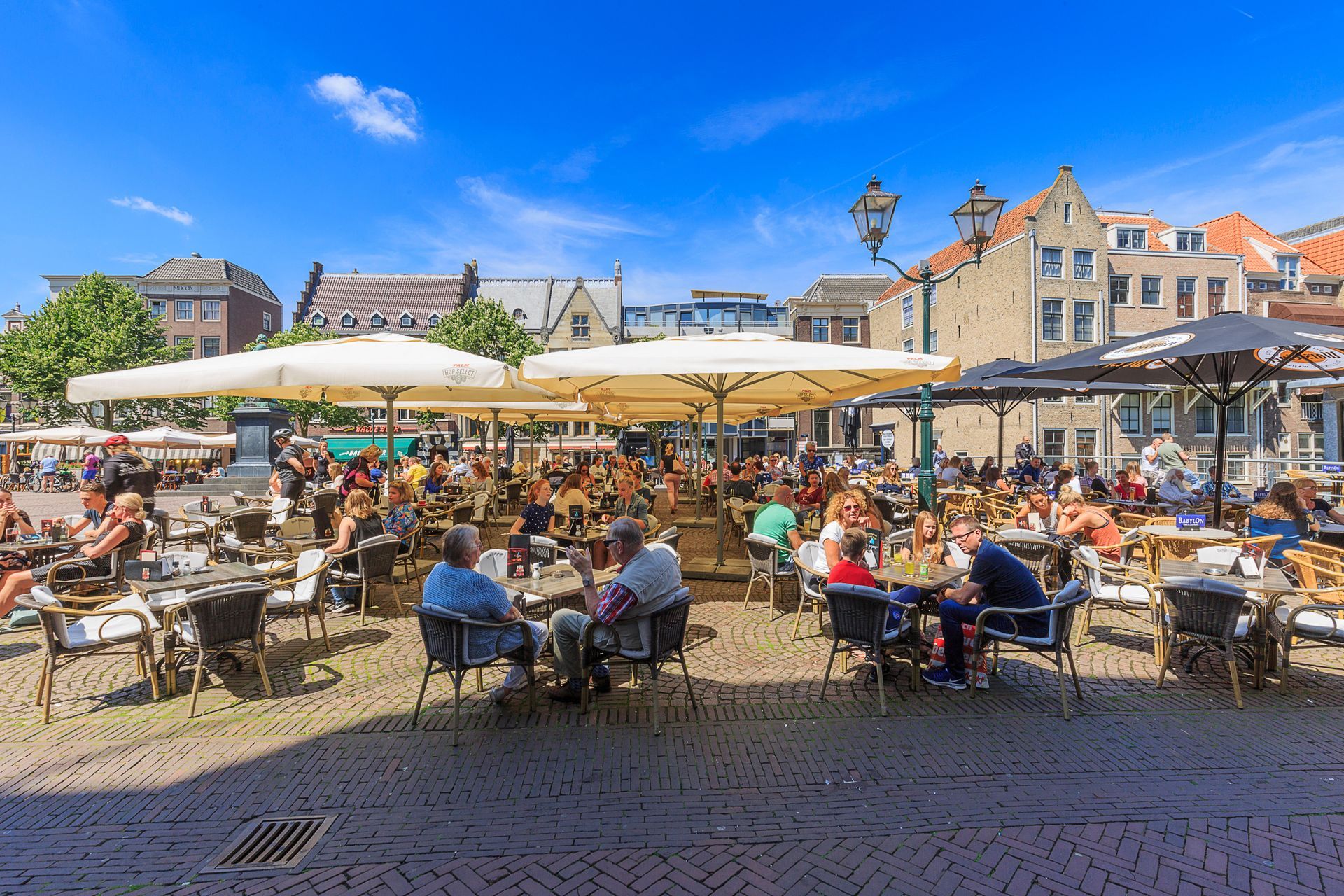Drukbezochte terrasjes op een zonnig stadsplein met historische gebouwen op de achtergrond.
