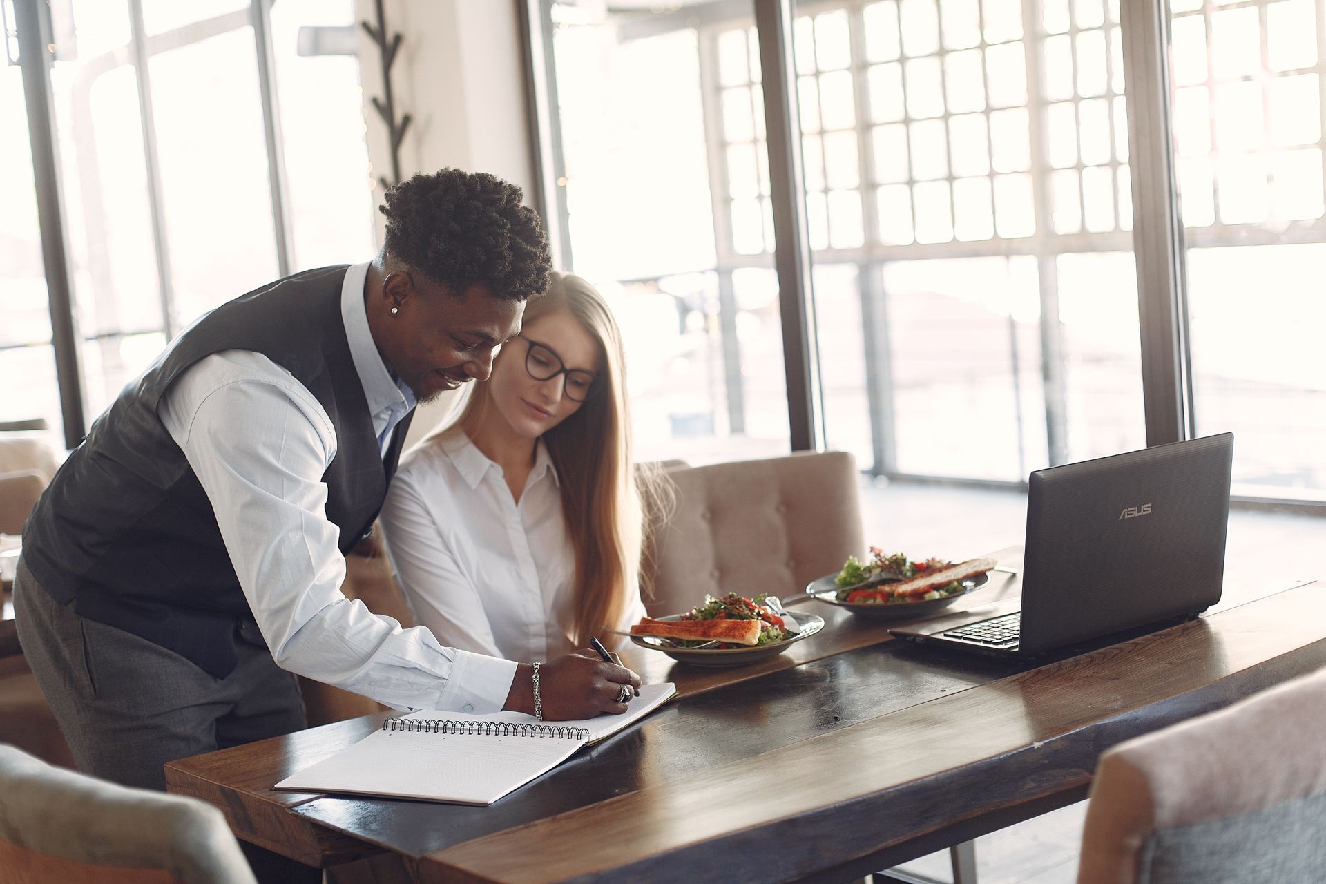 Twee collega's bekijken documenten aan een tafel met een laptop en wat te eten in een lichte kantoor
