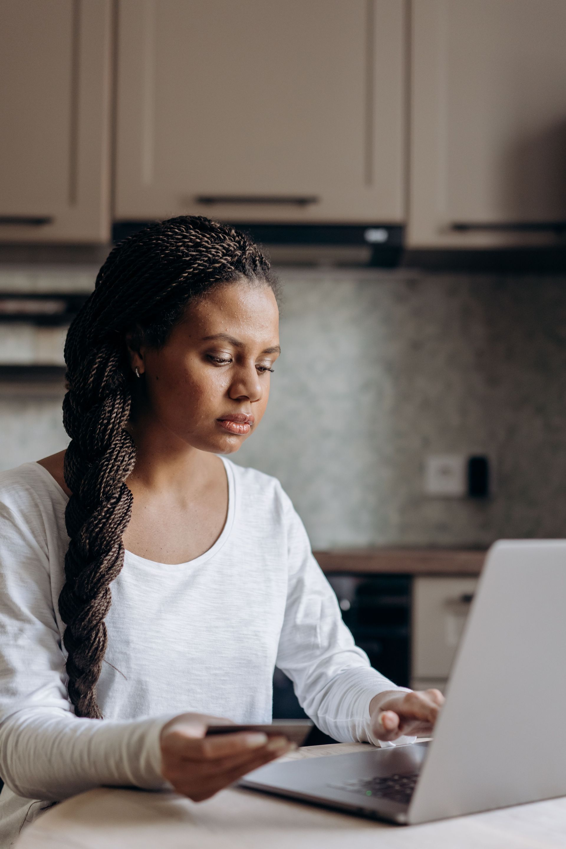 Person working on a laptop at a kitchen counter, holding a phone in a softly lit home kitchen.