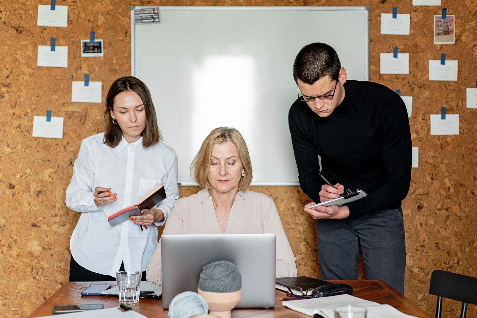 Three coworkers discussing paperwork around a laptop in a meeting room with a whiteboard.