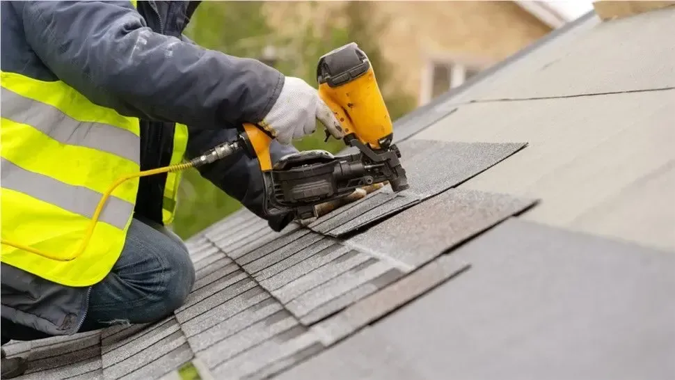Worker installing roof shingles with a nail gun on a sloped roof, wearing a high-visibility vest.