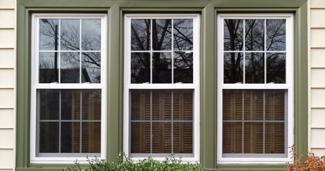 Three white-trimmed windows on a beige house exterior with green shutters and blinds inside
