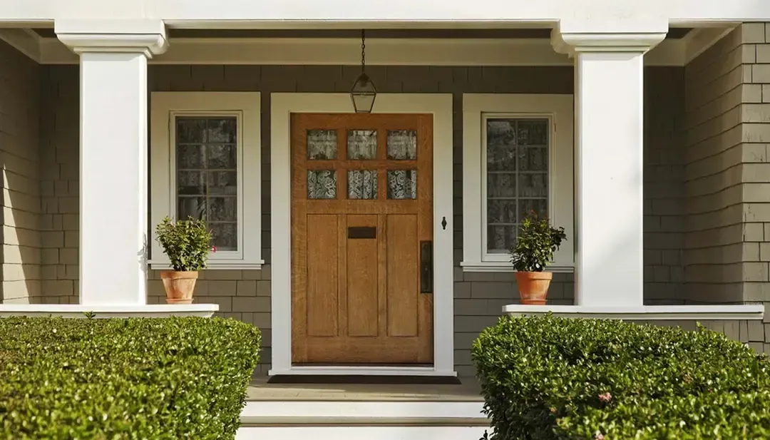 Front porch with a wooden front door, white columns, and potted plants, framed by trimmed hedges.