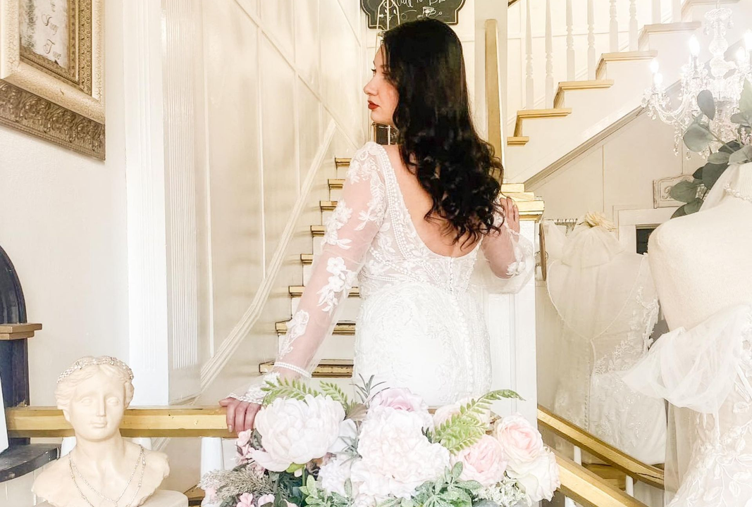 Bride in white lace dress, holding bouquet, smiles in front of a white building on a green lawn.