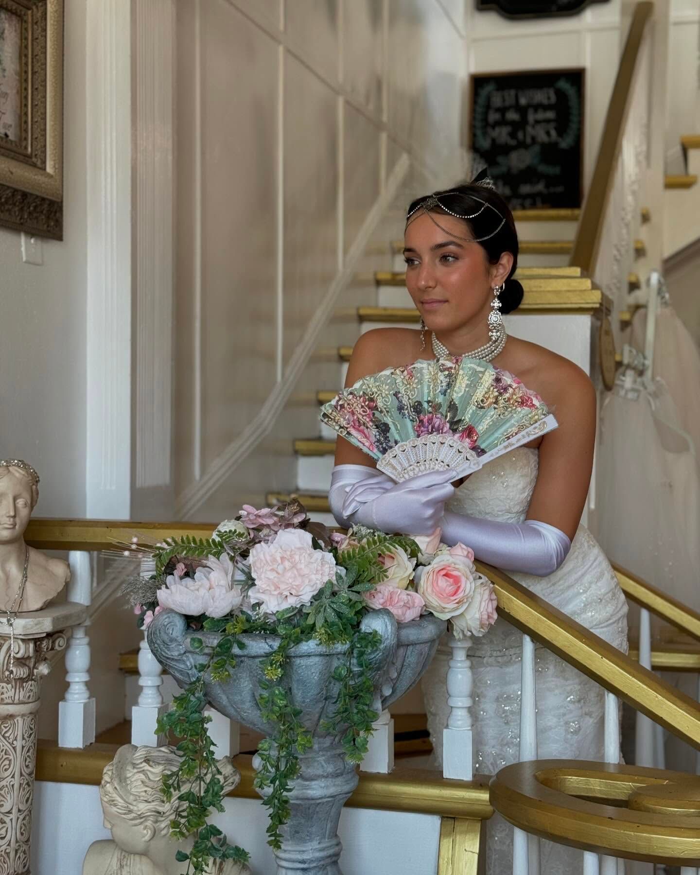 Woman in a flapper dress poses on stairs, holding a fan. Flowers in a large urn.