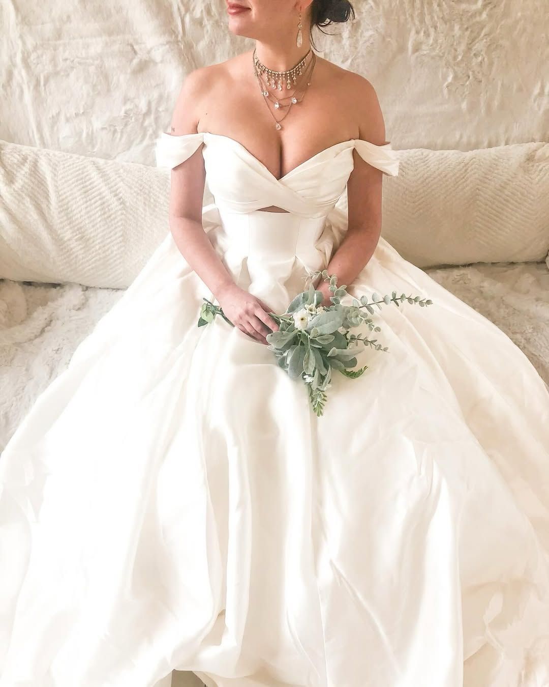 Woman in white off-the-shoulder wedding gown, holding bouquet, seated indoors.