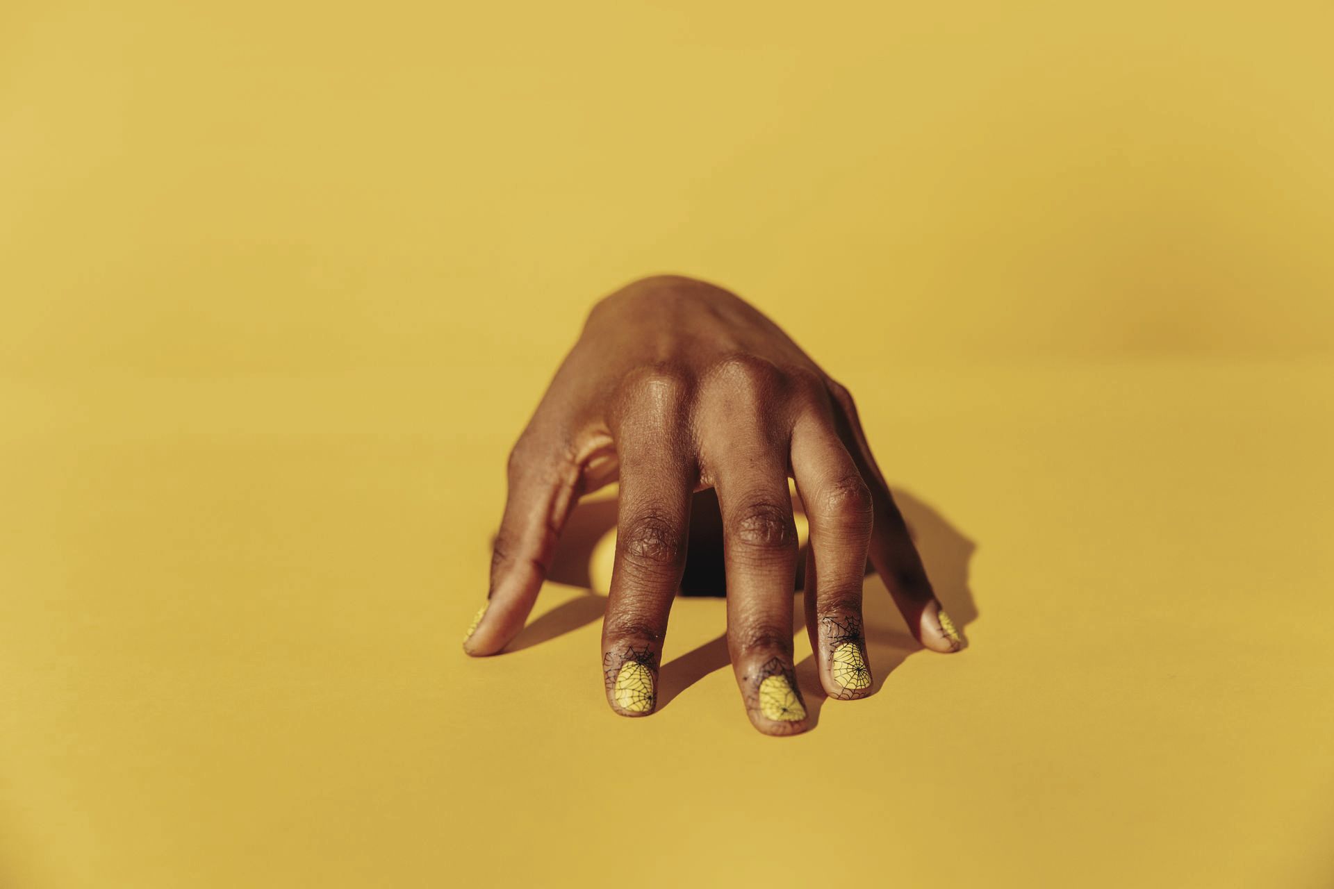 Hand with long gold-painted nails reaching up from a yellow surface against a yellow background
