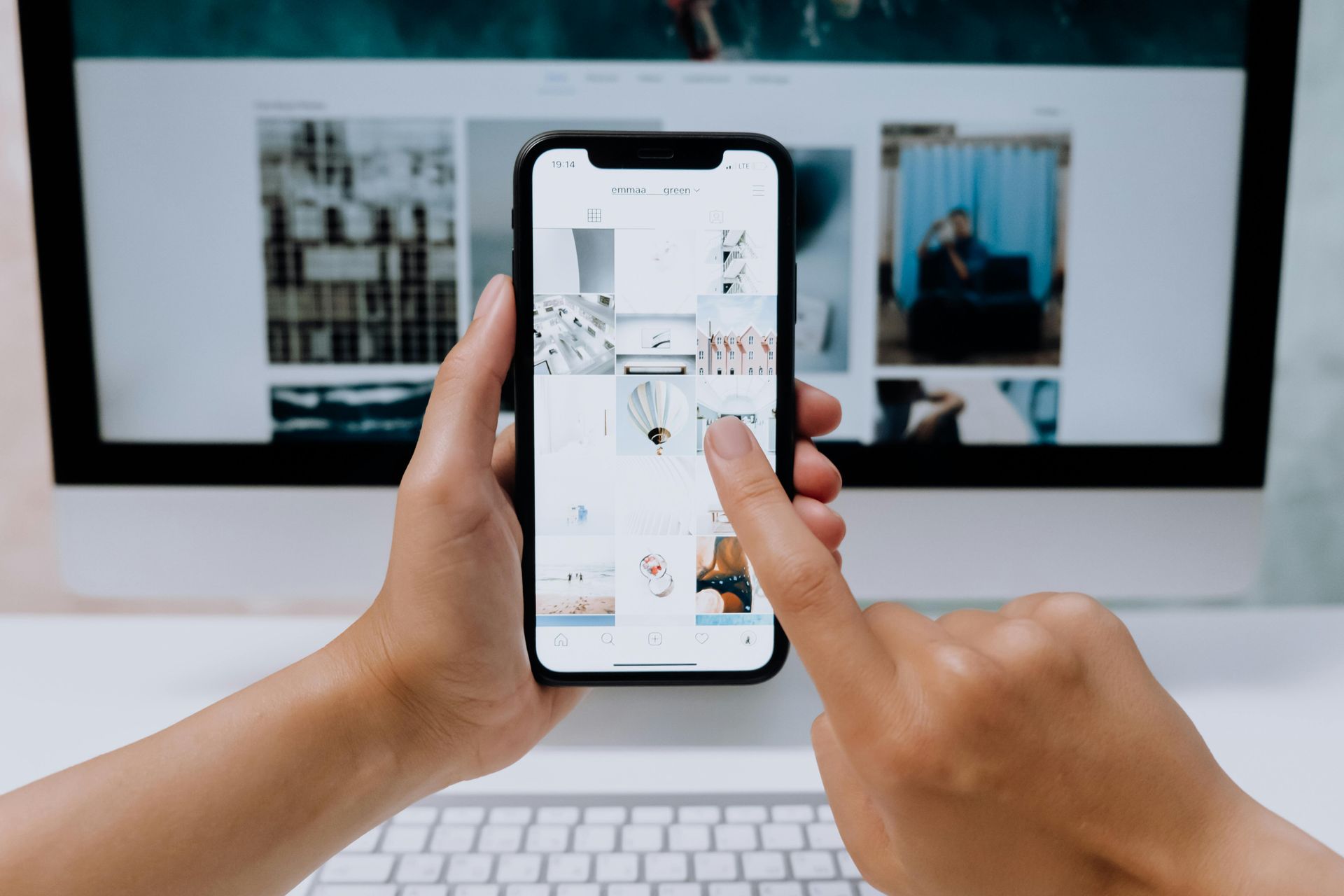 Hands holding a smartphone displaying an image grid, positioned in front of a computer screen showing similar content.