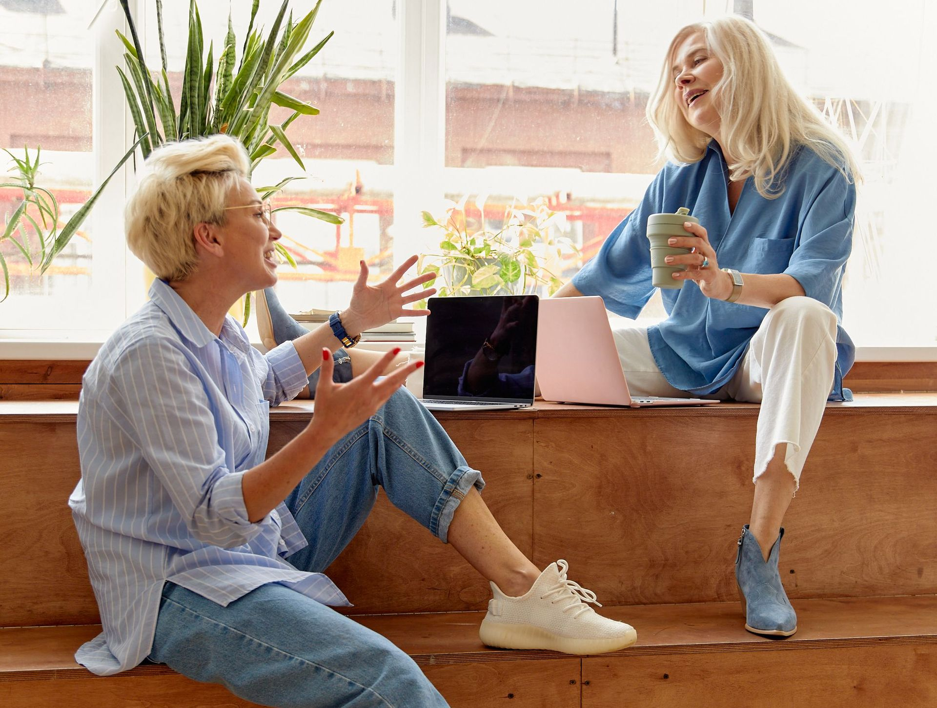Two people chatting by a window with a laptop and coffee in a bright office lounge