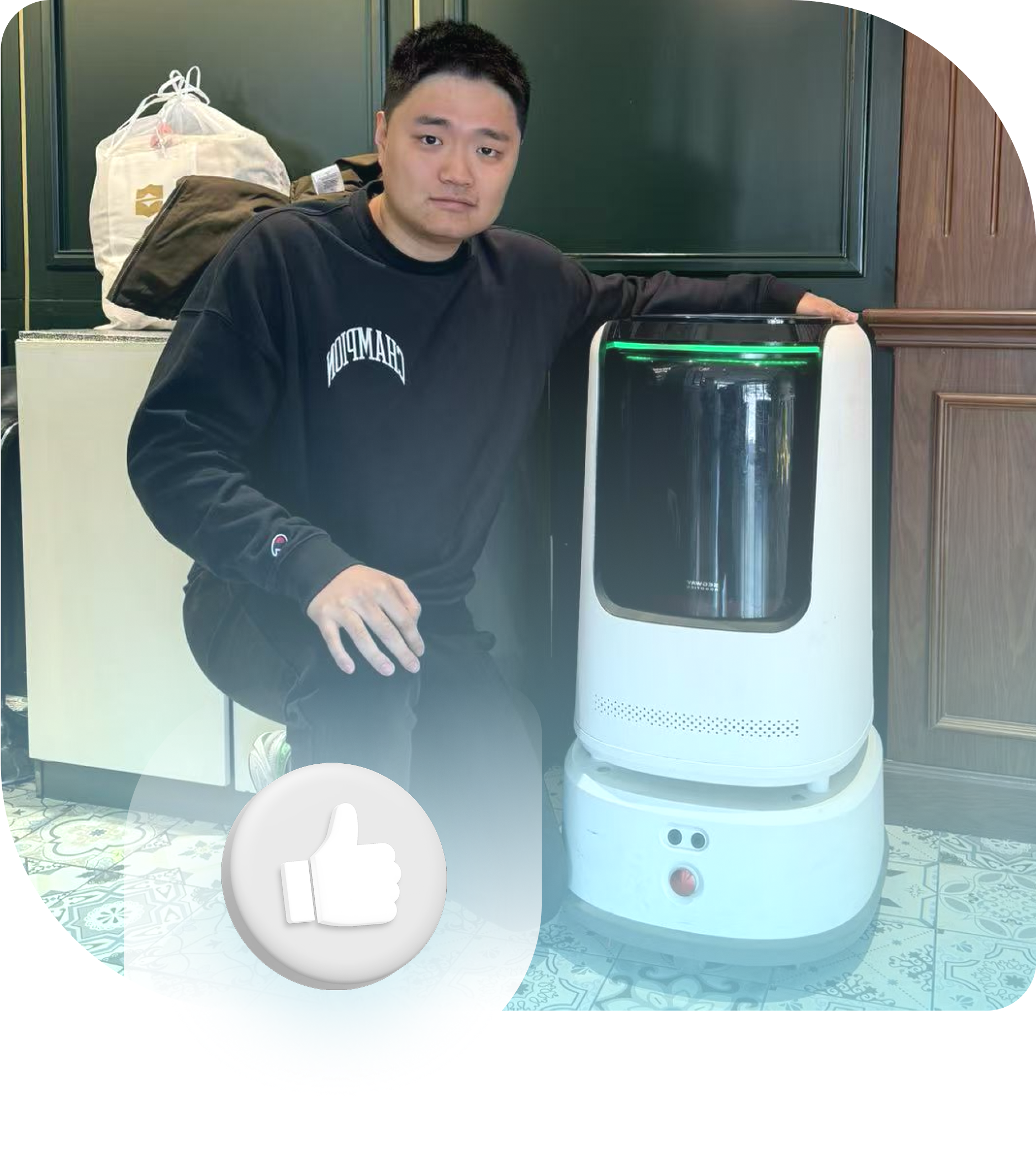 Man kneels next to a white robot. He's wearing black and smiles. Interior setting.