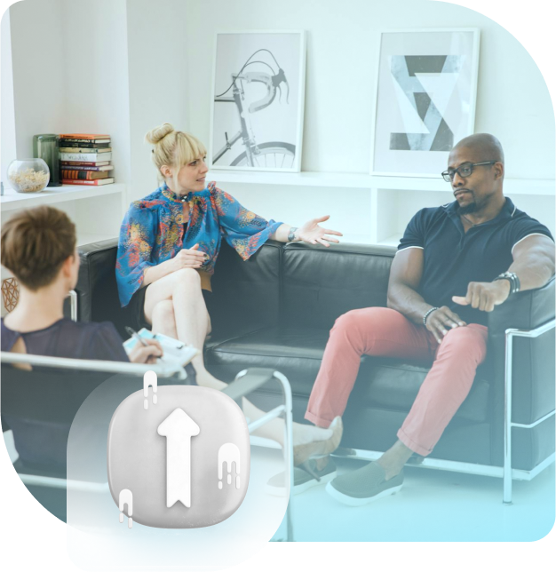 People in therapy session; woman gestures, man listens on couch, another sits on the side.