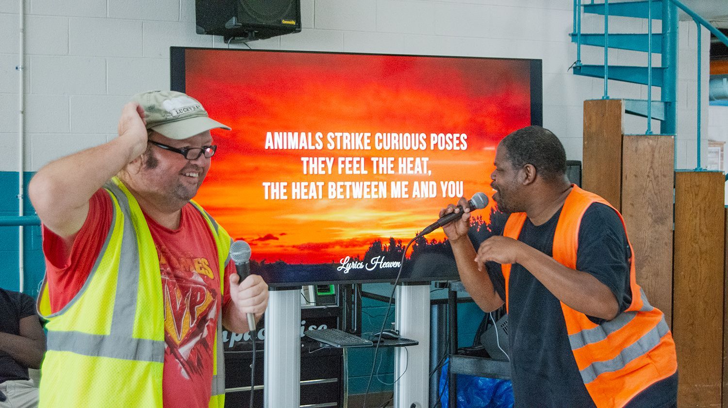 Two men wearing reflective vests performing during a karaoke event in the OGC cafeteria.