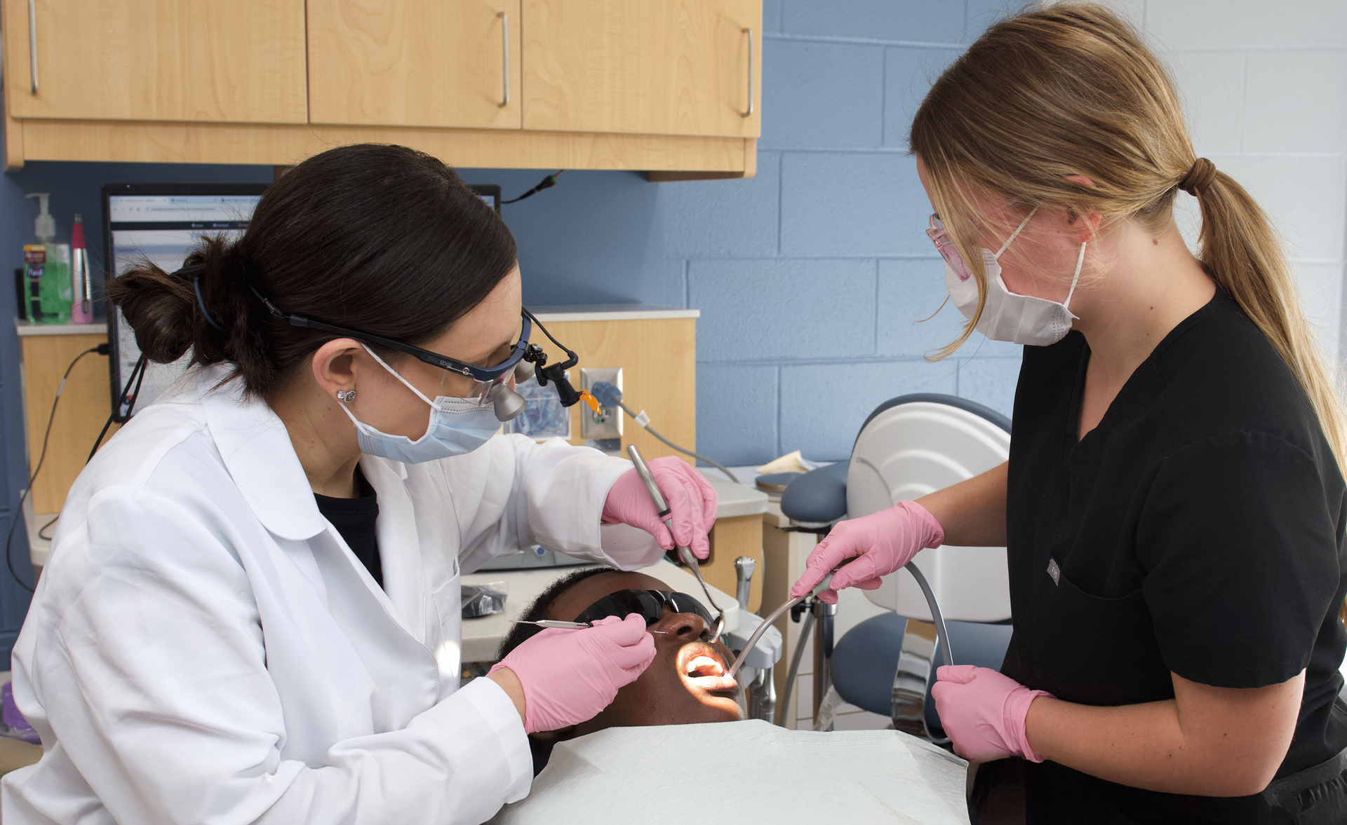 A man in a patient's chair in the dental clinic, with two dentists wearing gloves and masks grip dental tools.
