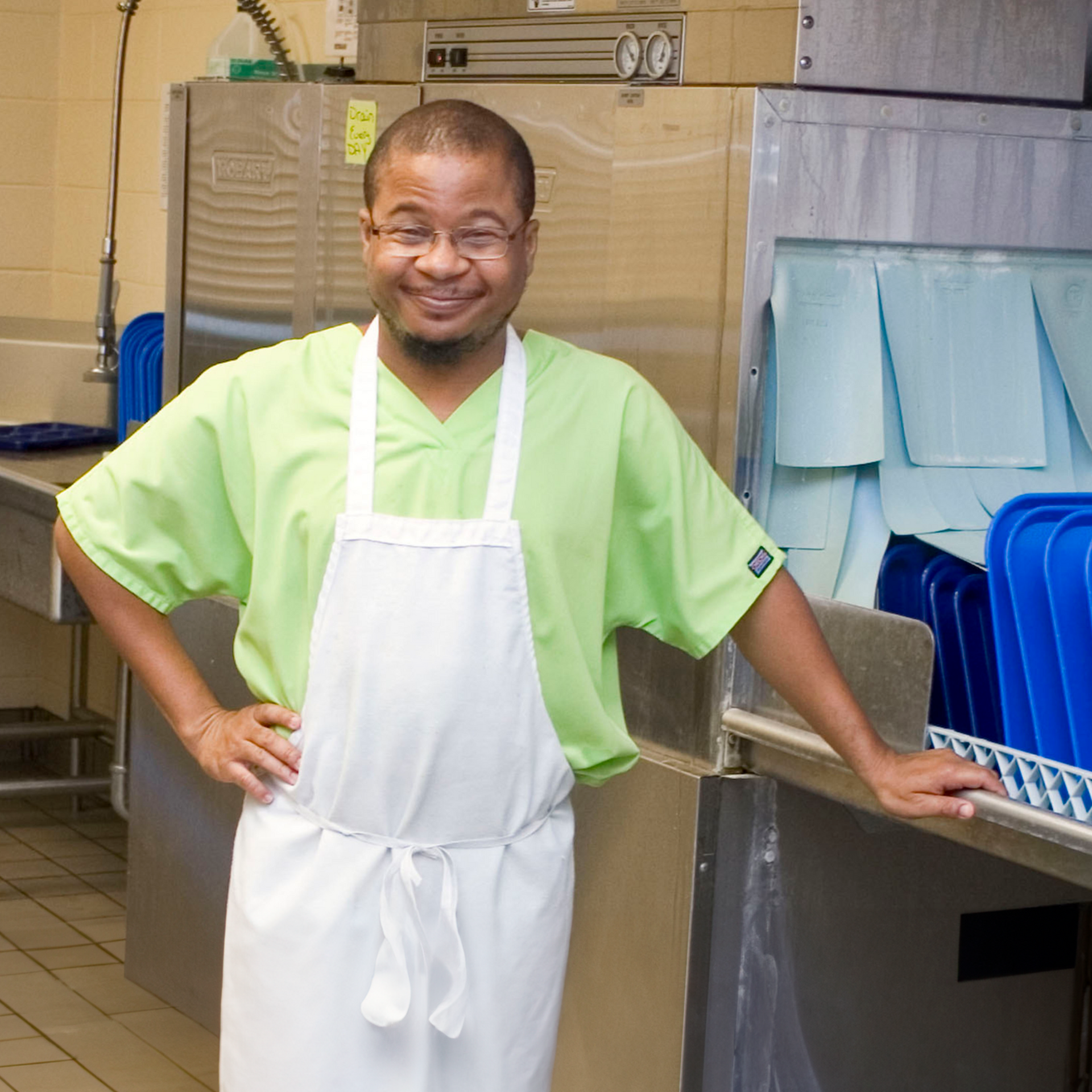 A man wearing glasses leans on a metal kitchen sink counter, posing and smiling in the middle of his work day.