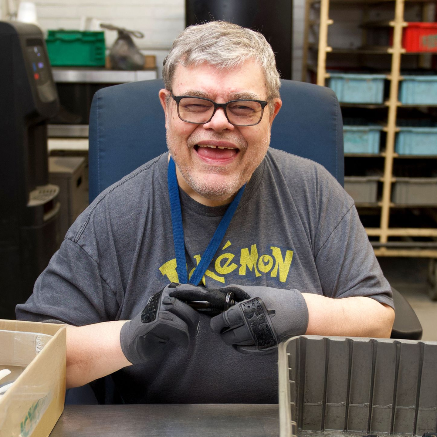 A man in a Pokemon shirt smiles big while handling metal pieces as part of his sorting job at Komatsu.