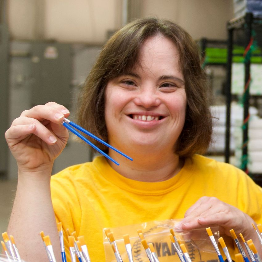 A woman smiles, holding paintbrushes while in the process of sorting them as part of her job at Kids Art Box.