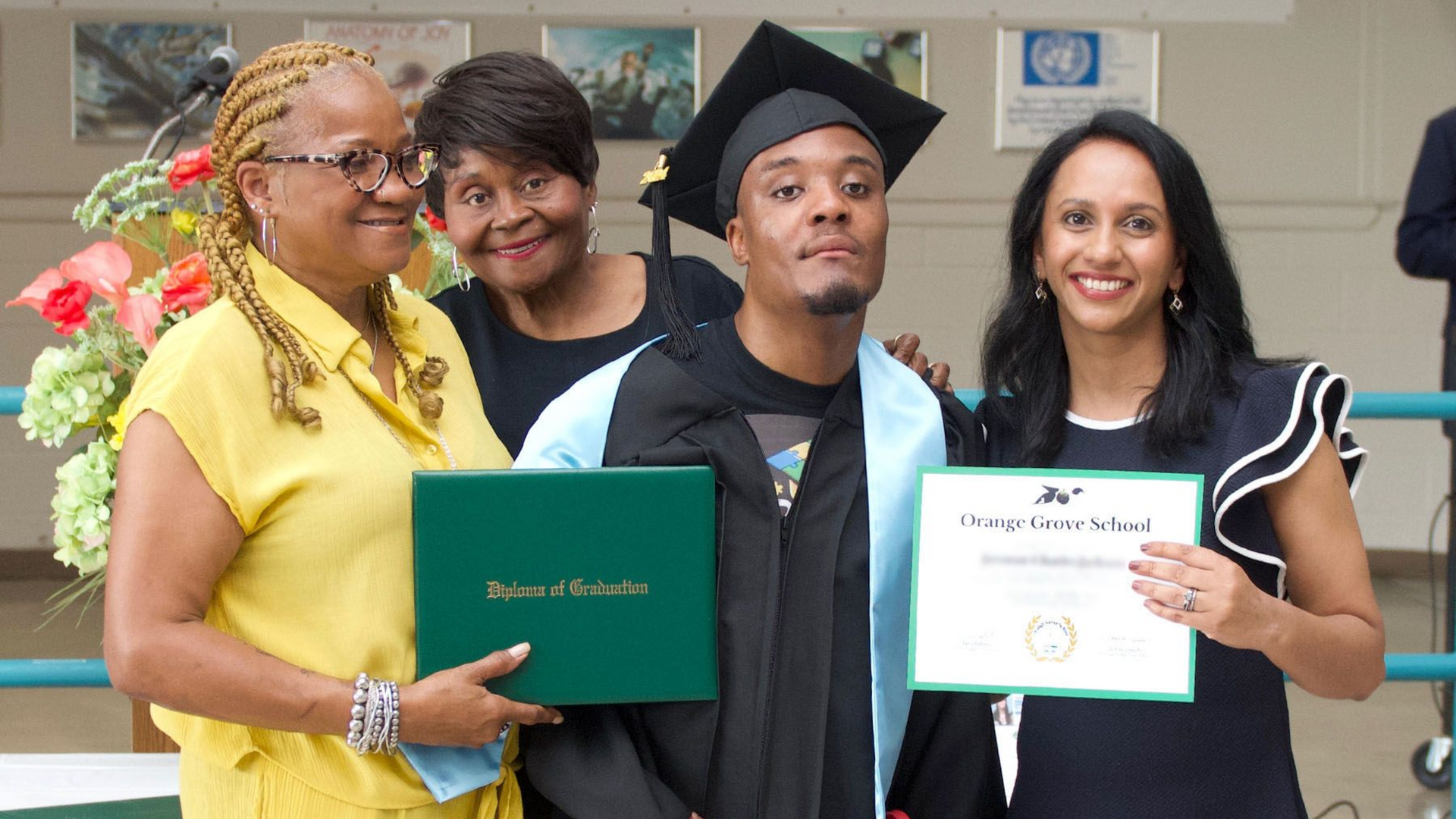 A group photo of a graduating male student of Orange Grove School, wearing a cap and gown, receiving his diploma.