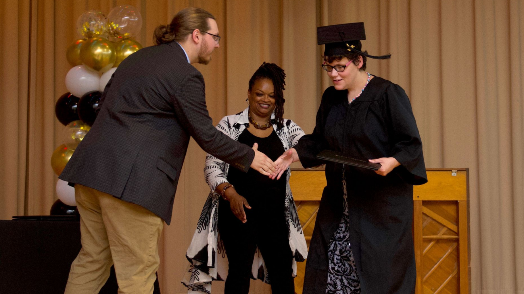 A man in a suit shakes the hand of a woman supported by Orange Grove wearing a cap and gown during a graduation ceremony.