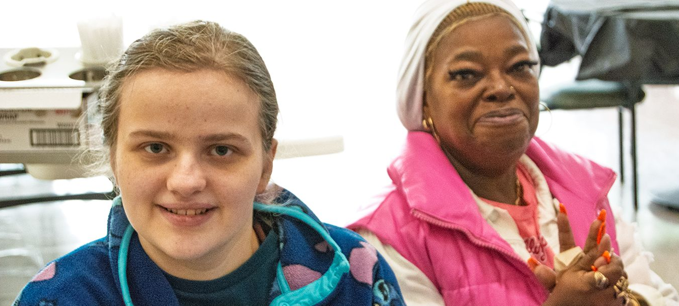 A female student of Orange Grove School smiles, wearing a fuzzy blue sweater, seated next to a female staff member.